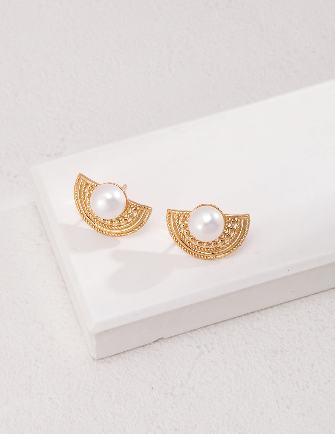 Gold-tone semicircular stud earrings with white faux pearls and intricate filigree detailing, displayed on a white platform